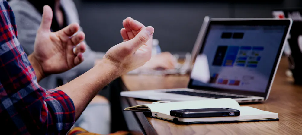 Dos personas conversando en una mesa de trabajo junto a un portátil con datos en pantalla